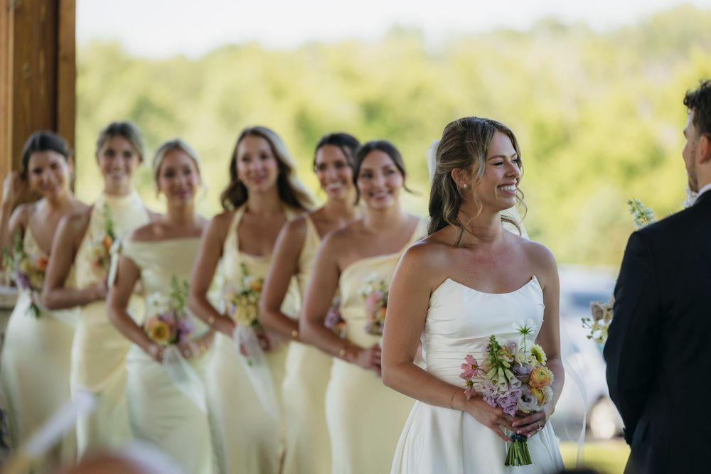 Smiling bride in strapless white gown holds bouquet with bridesmaids in butter yellow dresses lined up behind her at outdoor wedding.