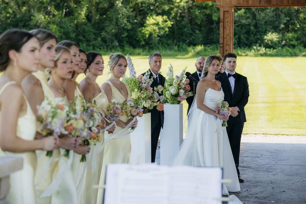 Bride and groom stand at outdoor wedding altar with bridesmaids in butter yellow dresses holding floral bouquets at Wisconsin vineyard