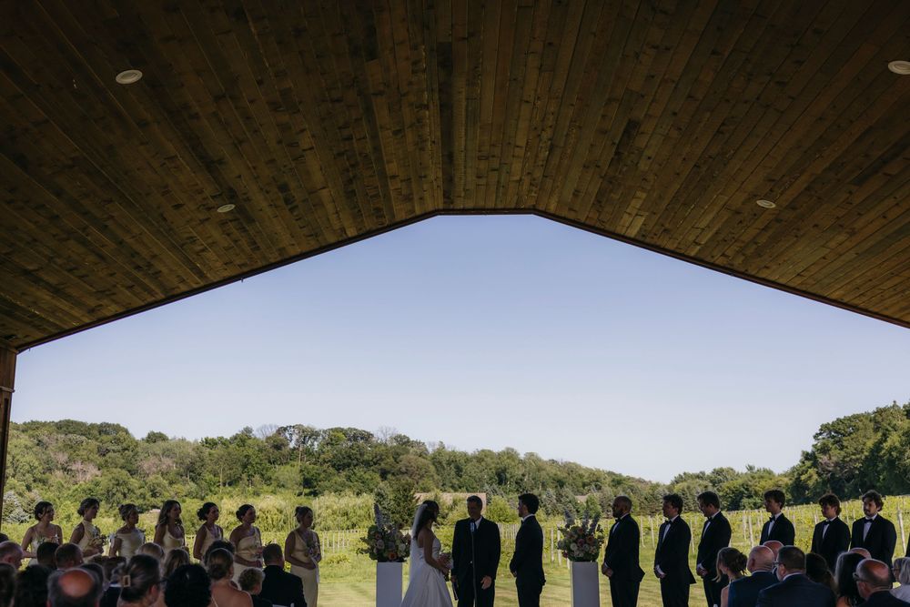 Outdoor wedding ceremony under wooden pavilion overlooking lush green field with guests and wedding party silhouetted at Wisconsin vineyard