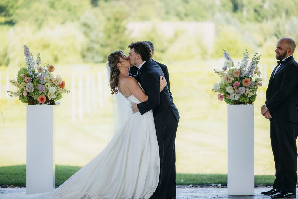 Bride and groom share first kiss at outdoor wedding ceremony with white pedestals and colorful floral arrangements at Wisconsin vinetyard