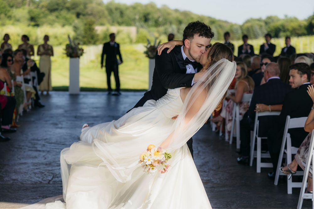 Groom dips bride in white gown during outdoor wedding ceremony kiss, guests seated in white chairs on sunny day in Wisconsin