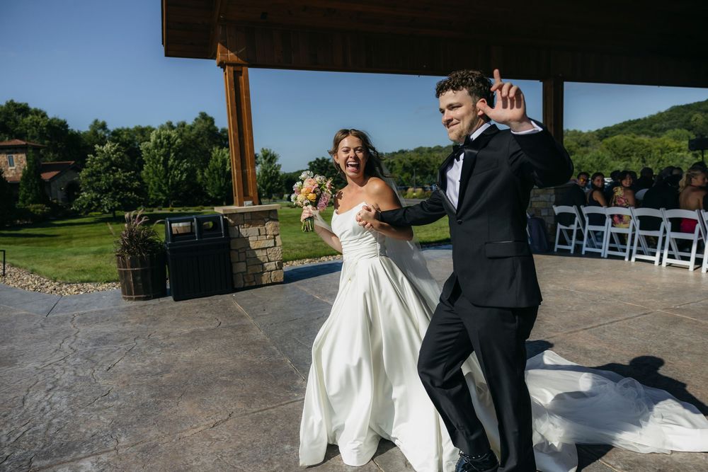 Joyful newlyweds in wedding attire celebrate their reception entrance at an outdoor Wisconsin venue with scenic green hills.