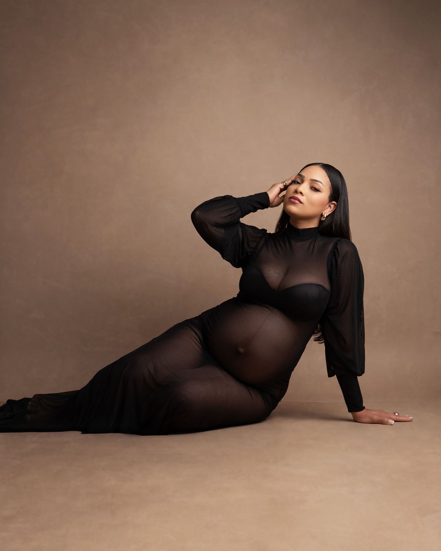 Pregnant woman in sheer black maternity gown poses gracefully on floor against warm brown studio backdrop.