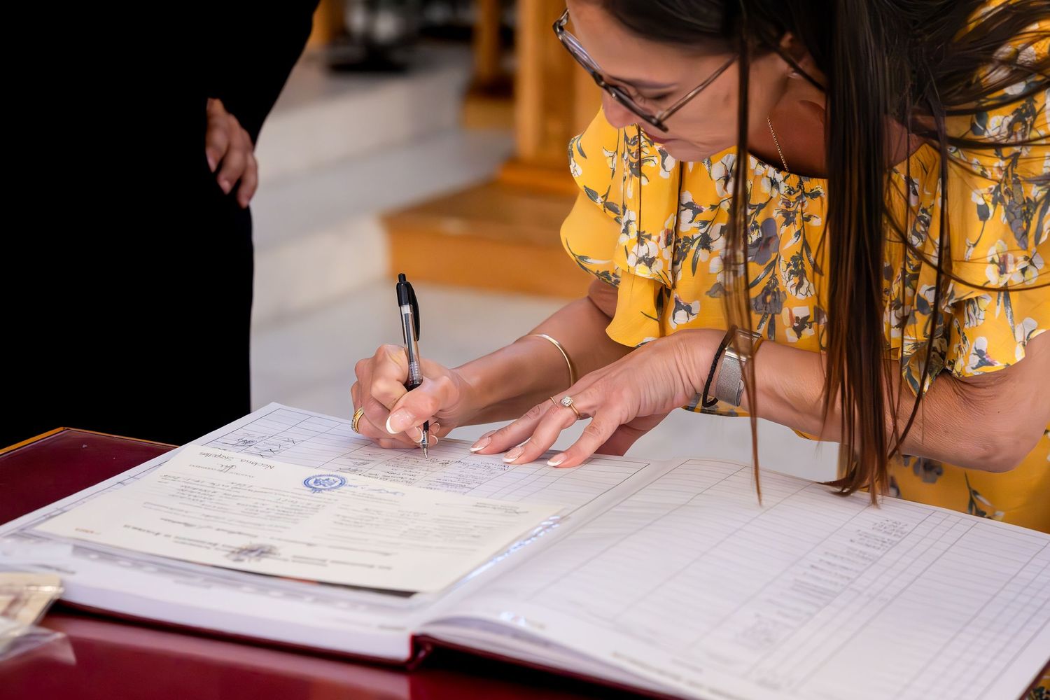 Woman in yellow floral dress signing official documents with a pen at a formal ceremony.