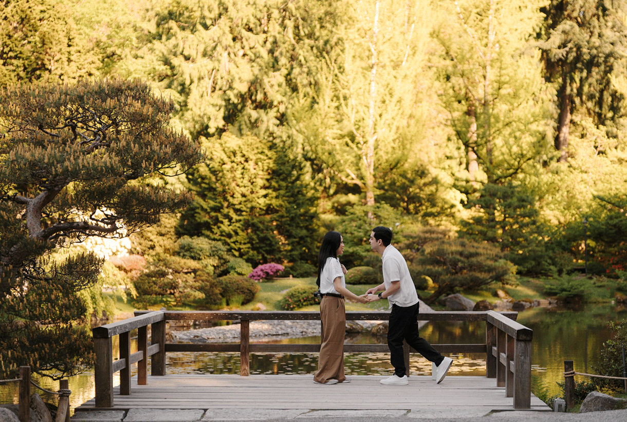 Two people stand on a wooden bridge in a lush Japanese garden with golden sunlight filtering through green trees.