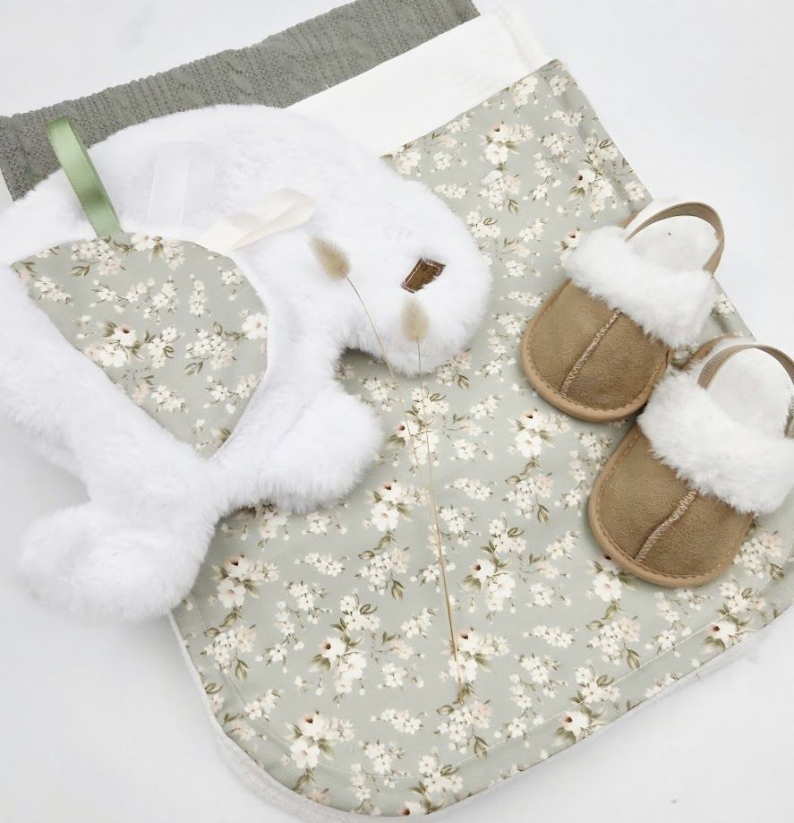 Sage green floral baby gift set with stuffed bunny, knit hat, and fur-trimmed sandals on white background.