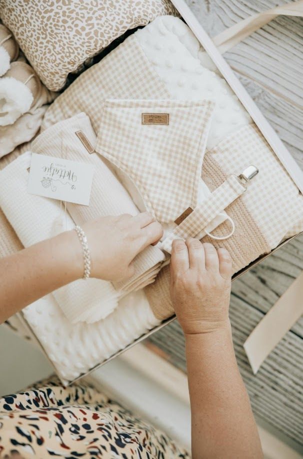 Hands arranging a neutral-toned baby gift set with gingham pouch, muslin blanket, and accessories in a wooden crate.