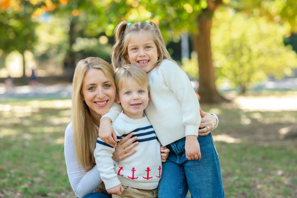 A smiling blonde mother poses outdoors with her two young children in a sunny park setting.
