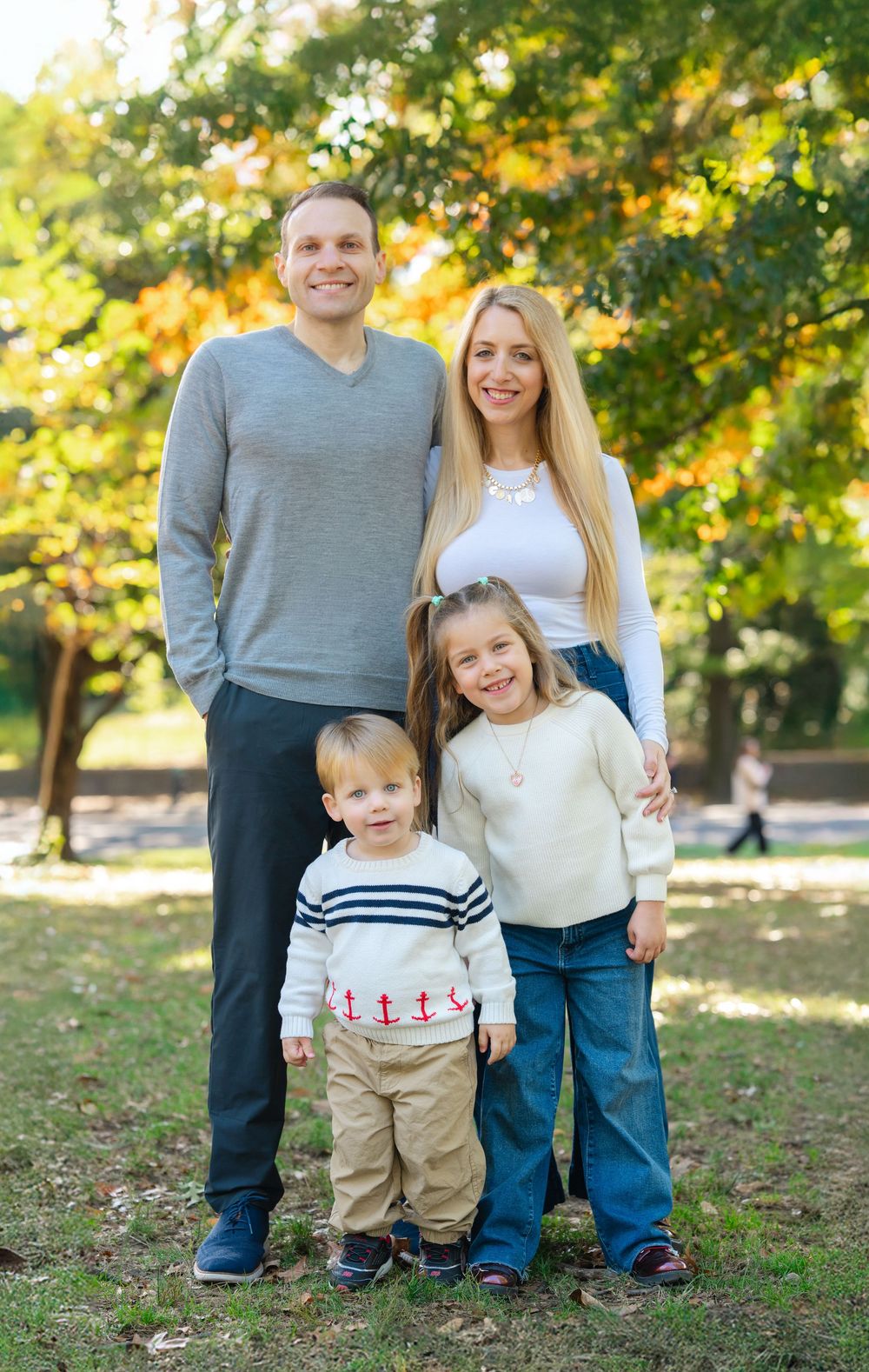 A happy family of four poses together in a sunny autumn park, parents standing behind their two young children.