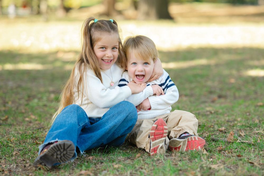 Two happy children sitting on grass in a park, smiling girl hugging laughing toddler boy outdoors.