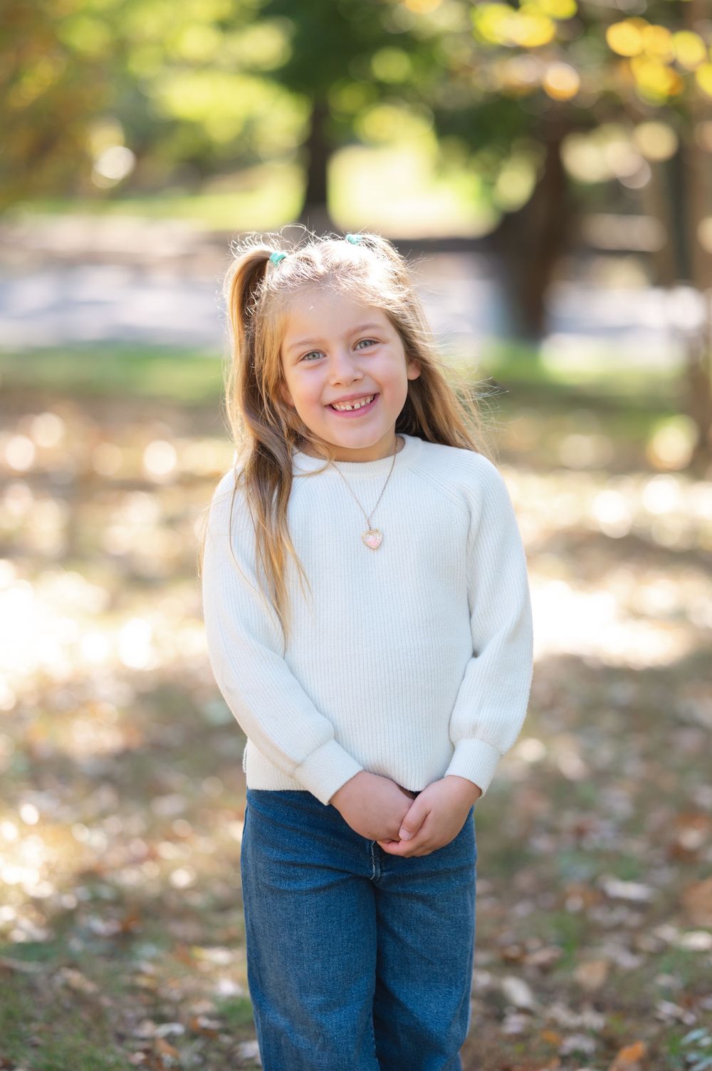 Smiling young girl in white sweater and jeans wearing heart necklace, standing outdoors in autumn park.