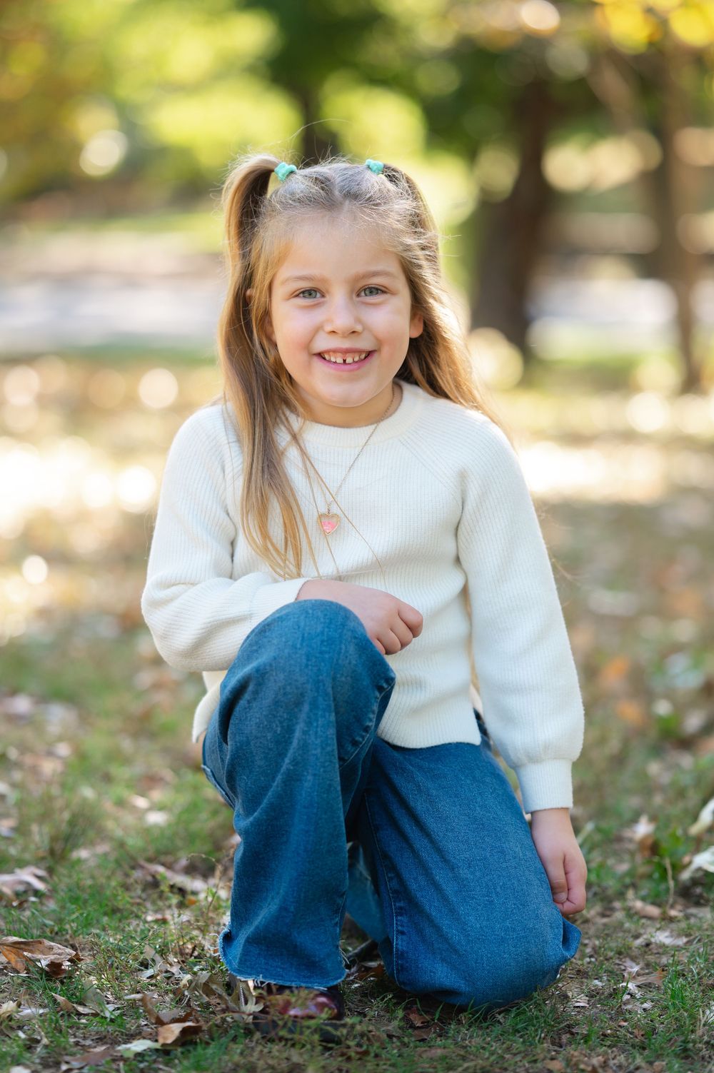 Young girl with pigtails in white sweater and jeans kneeling outdoors in autumn park, smiling brightly.