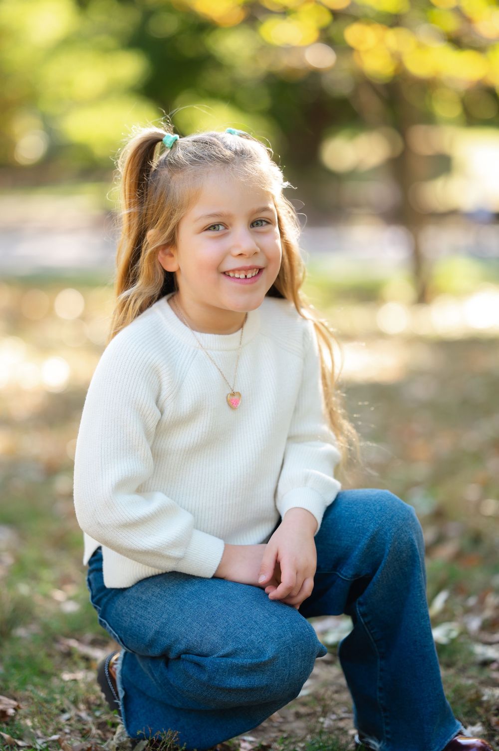 Smiling young girl with ponytail wearing white sweater and jeans sitting outdoors in a sunny park setting.