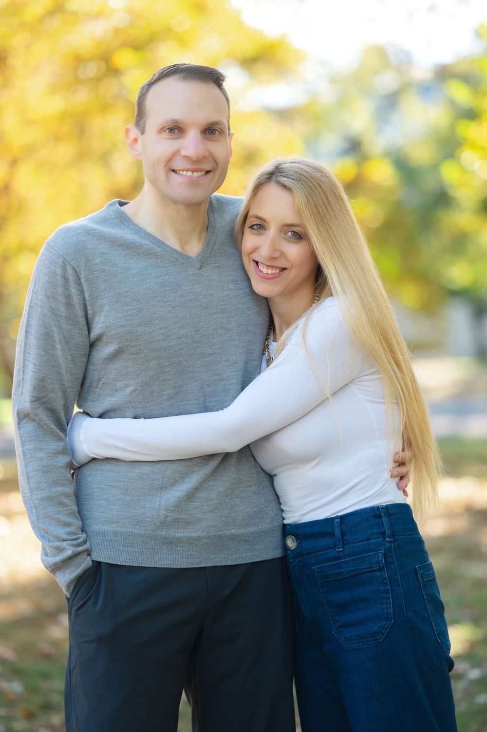 Smiling couple embracing outdoors in autumn park, man in gray sweater and woman in white top and blue jeans.