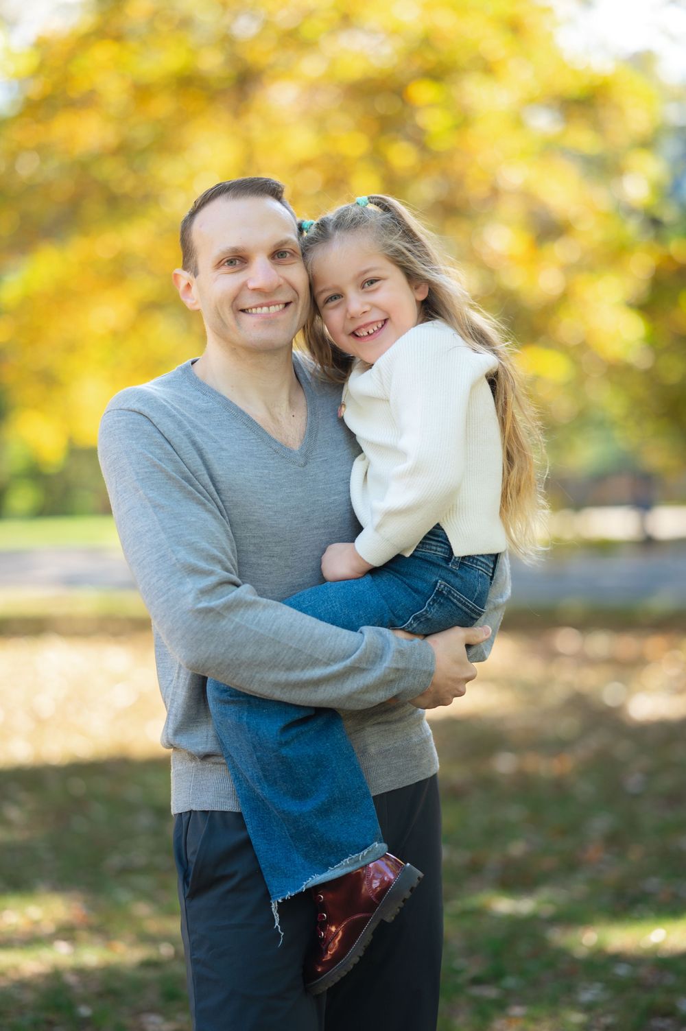 A smiling father holds his young daughter in a sunny autumn park with golden foliage in the background.