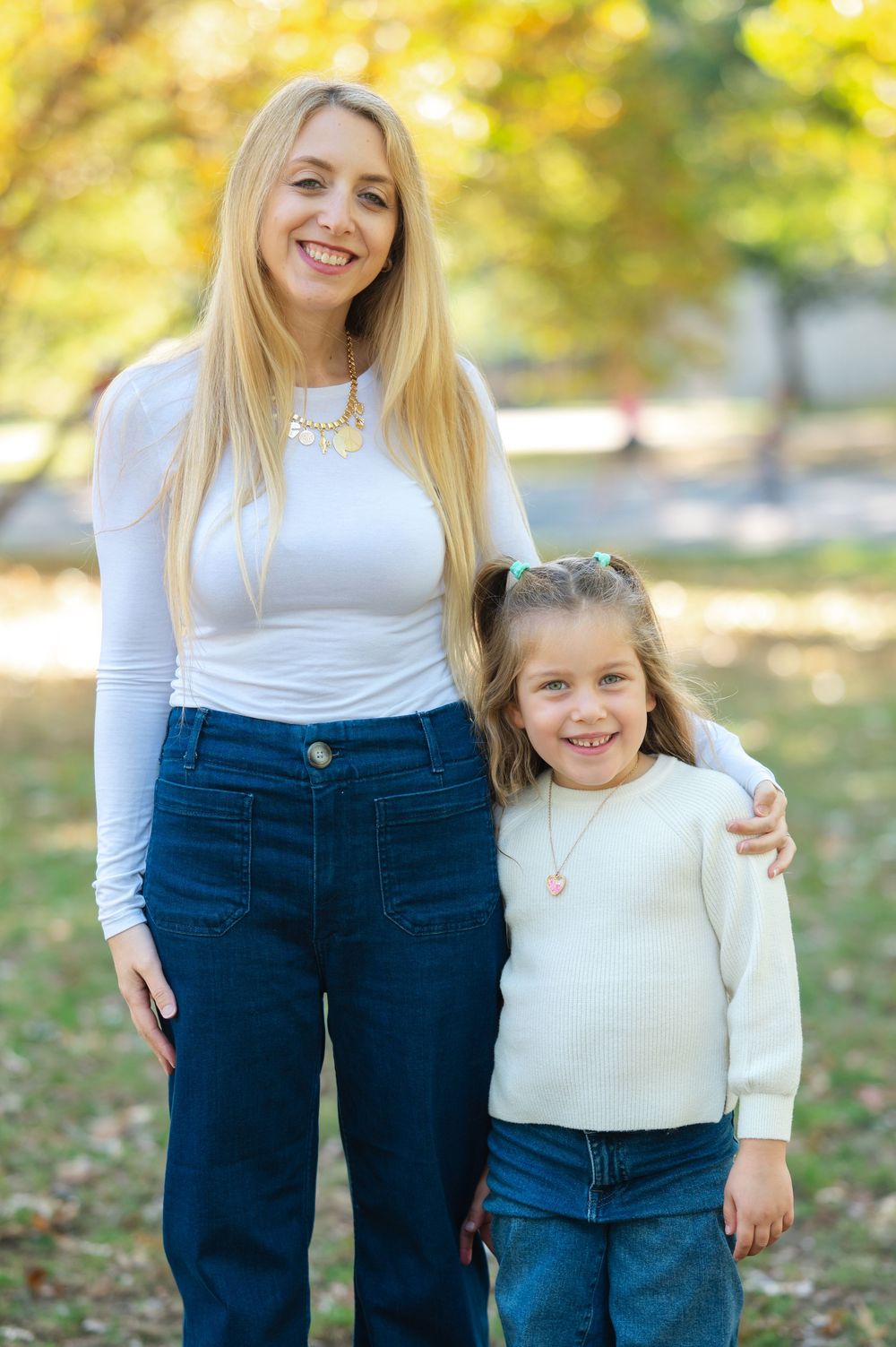 Smiling blonde woman and young girl in matching white tops and jeans posing together outdoors in autumn park.