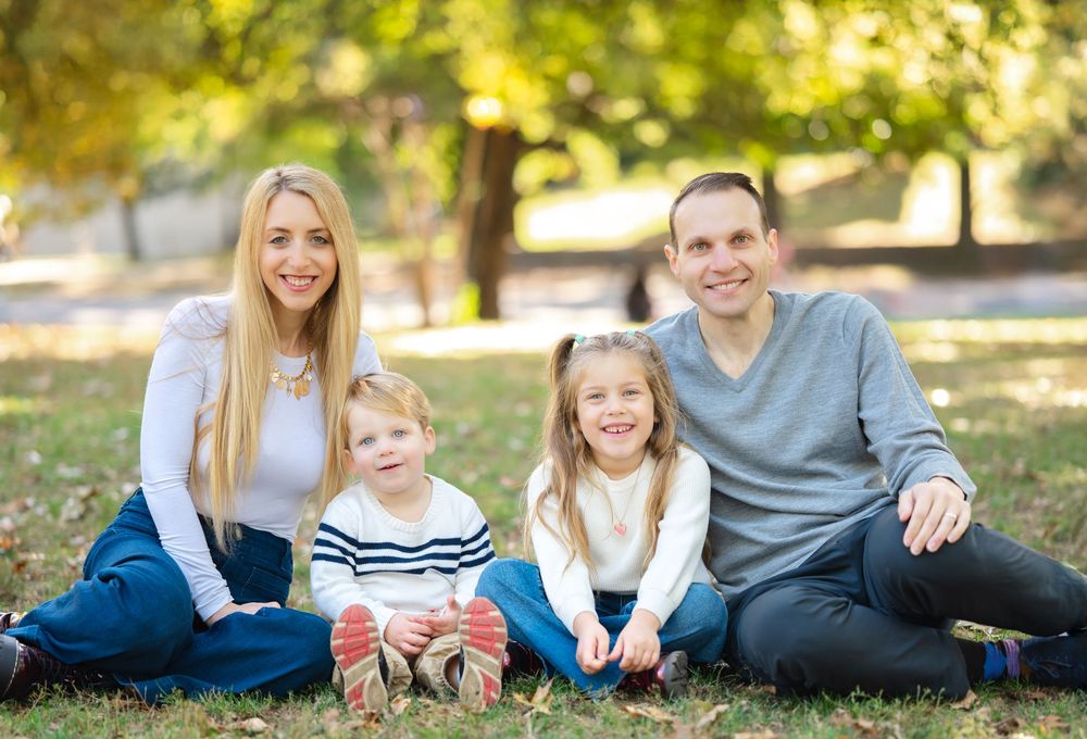 A happy family of four poses together on grass in a sunlit park during fall season.