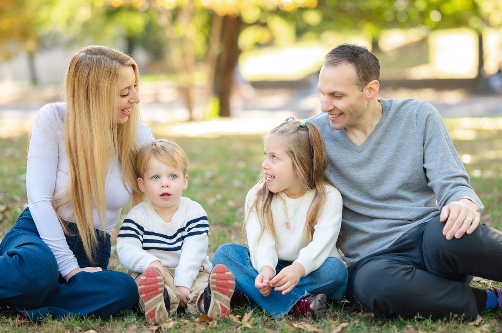 Happy family of four sitting together on grass in a sunny autumn park, smiling and bonding.