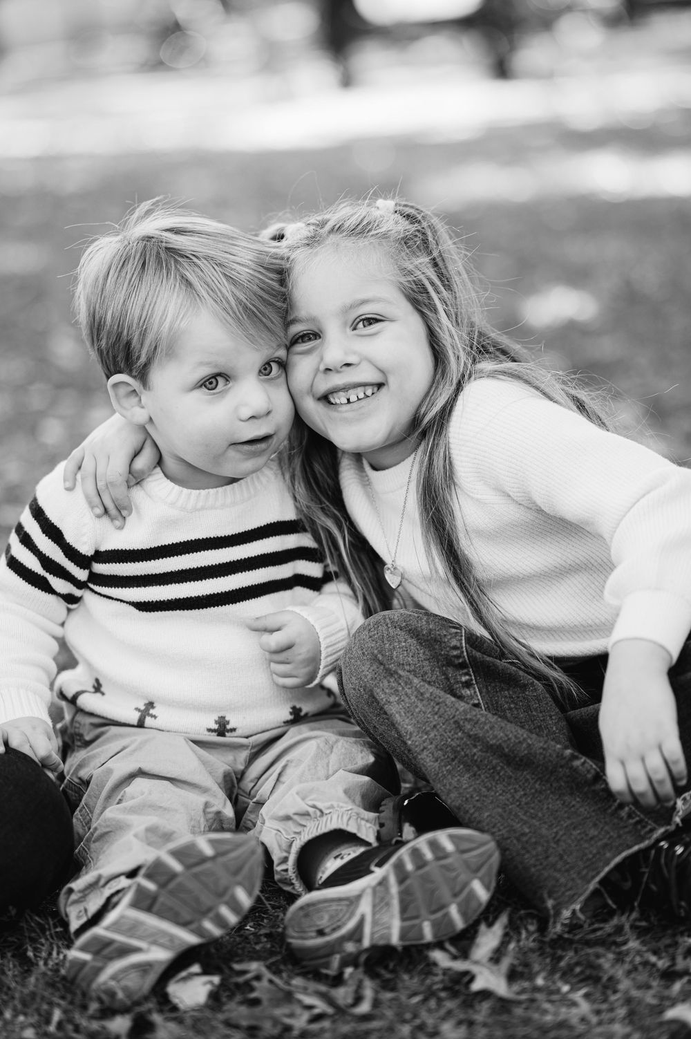 Black and white photo of two young siblings sitting outdoors, older girl smiling and hugging younger boy.