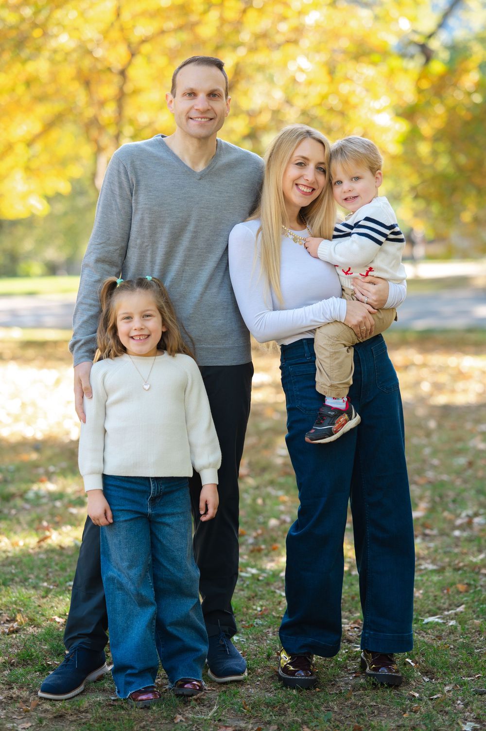 Smiling family of four posing together in a colorful autumn park, mother holding young toddler boy.
