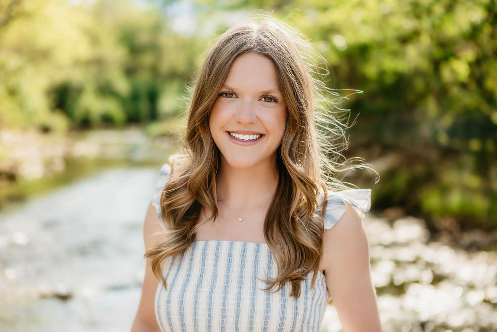 Smiling young woman with long brown hair wearing a striped top, posing outdoors near a creek.