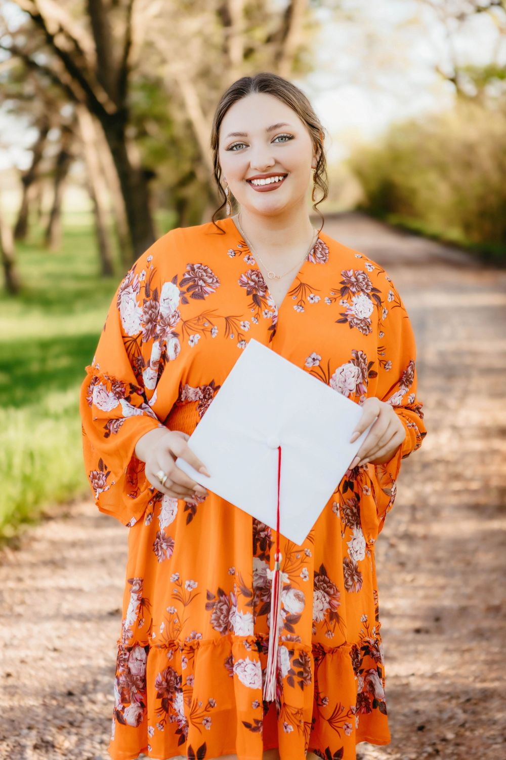 Smiling graduate in orange floral dress holding white cap with red tassel on a tree-lined path.