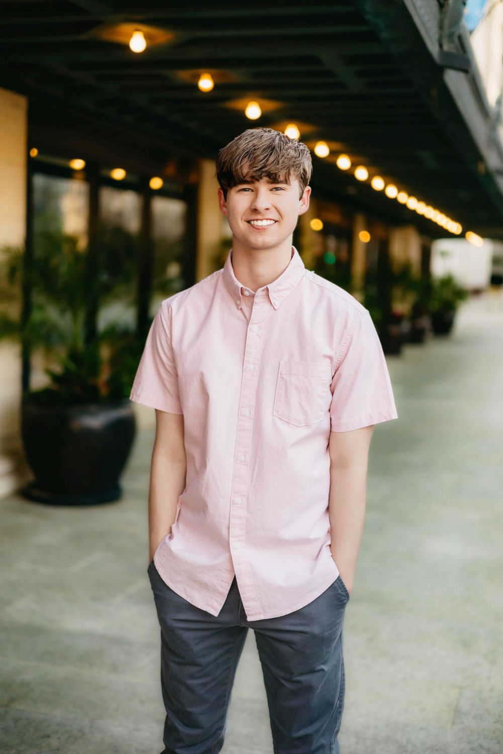 Young man smiling in pink short-sleeve button-up shirt standing outdoors under string lights.