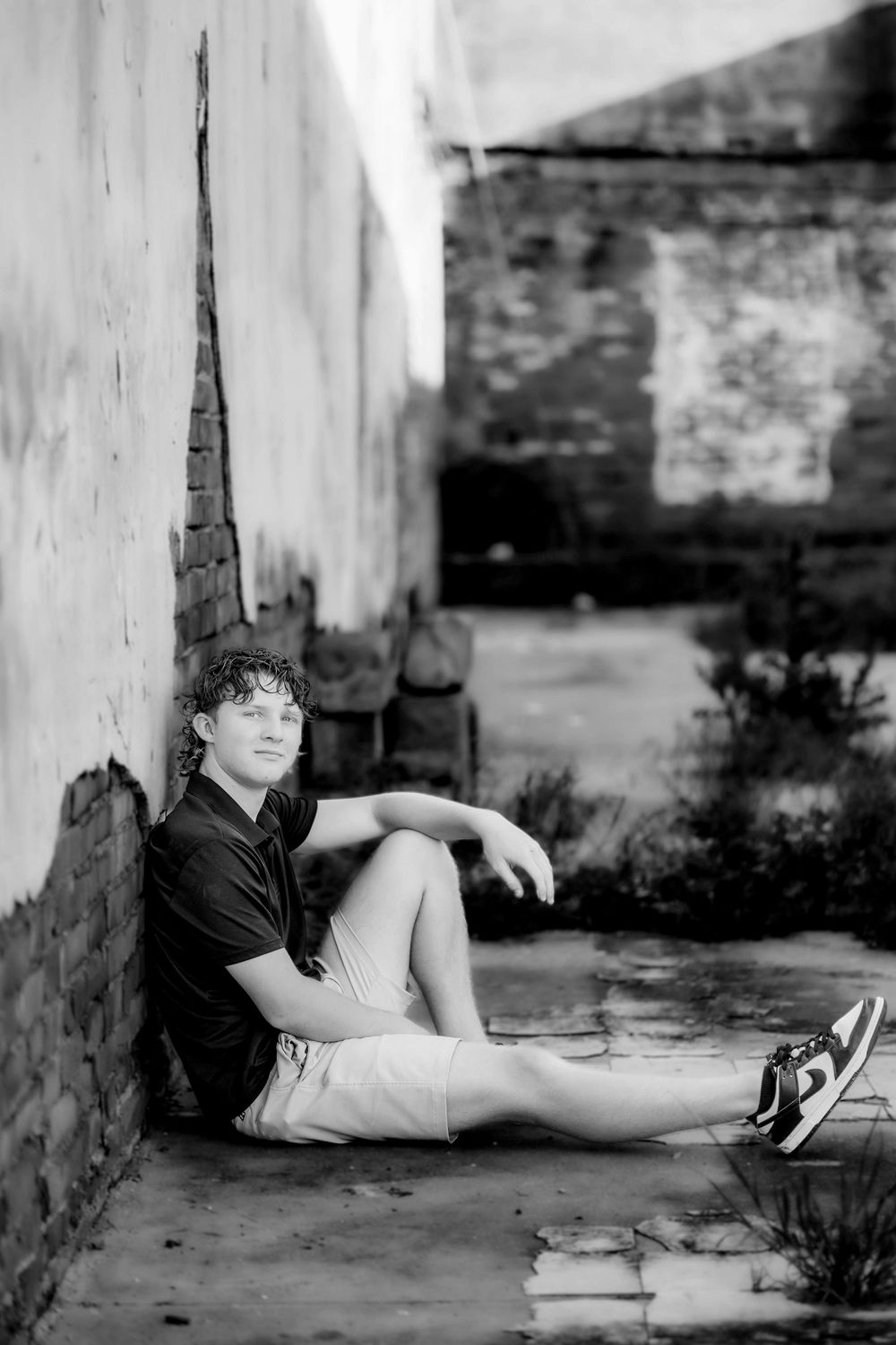 Young man in black polo and shorts sitting against a weathered brick wall in an abandoned building, black and white photo.