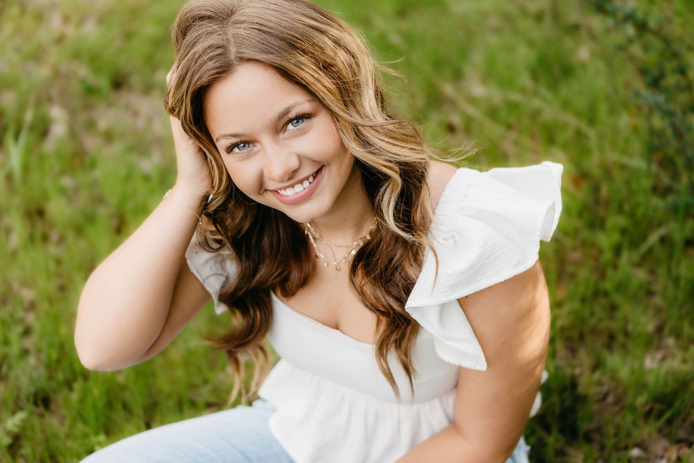Smiling young woman with wavy hair in white ruffled top sitting outdoors on green grass.