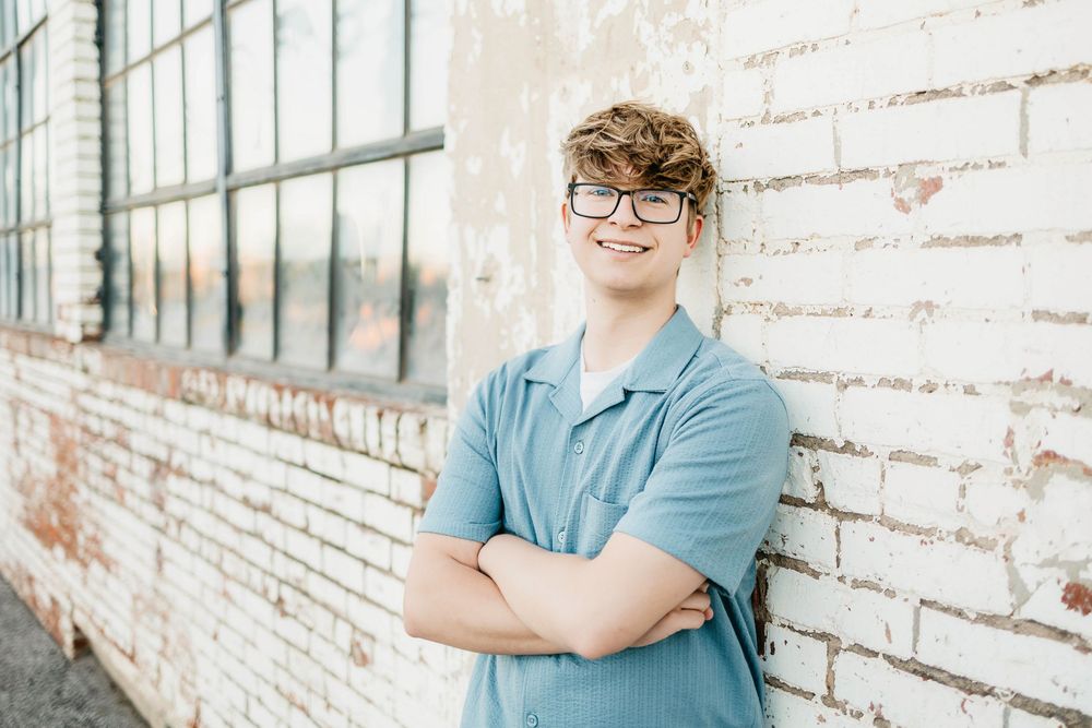 Smiling young man with glasses and crossed arms leaning against a white brick wall in a blue polo shirt.