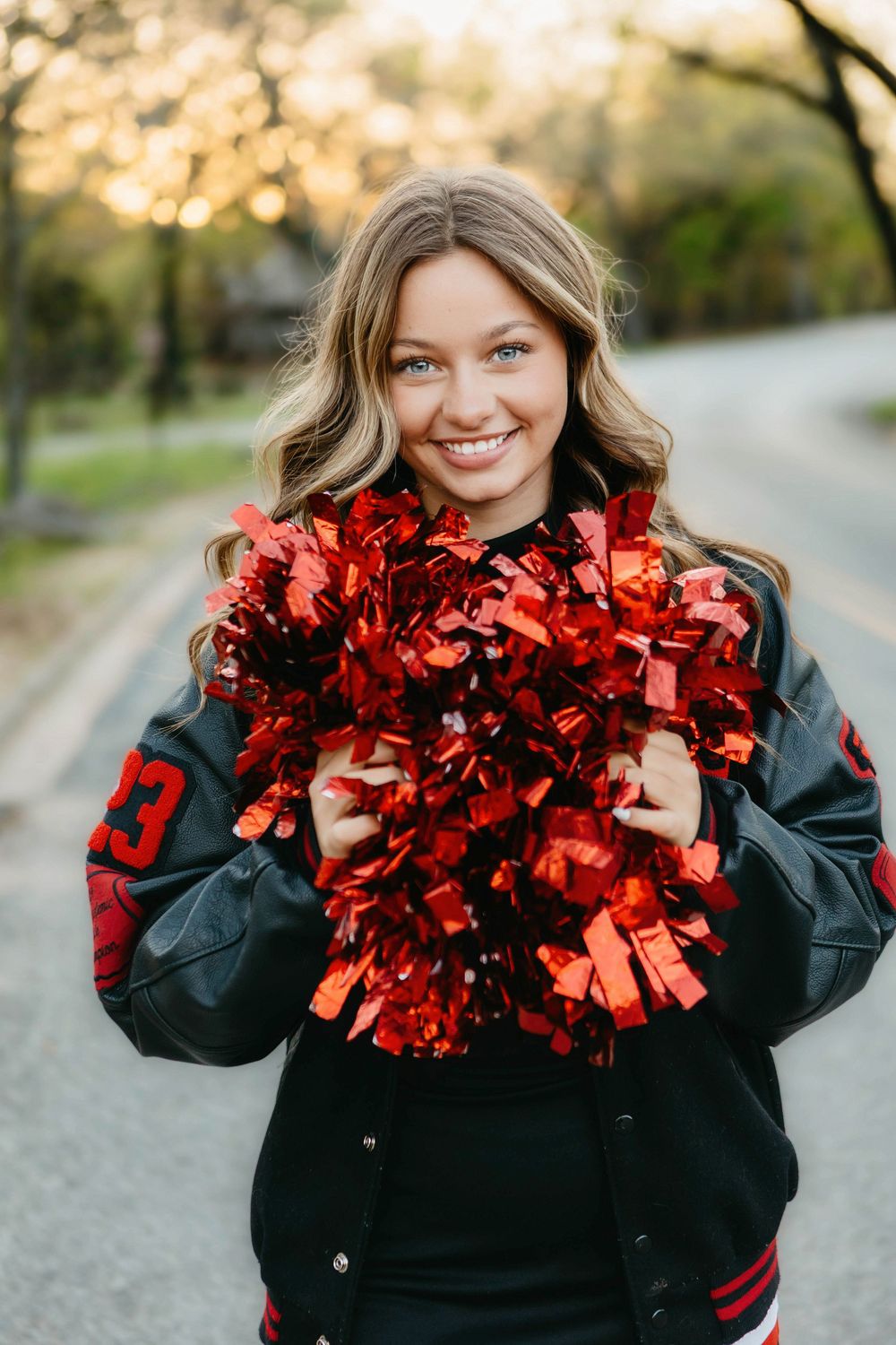 Smiling cheerleader holding red pom-poms wearing a black varsity jacket outdoors in warm golden light.