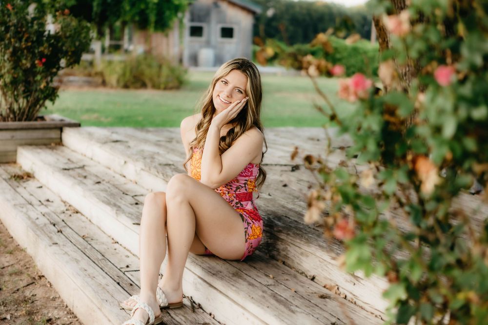 Young woman in floral romper sitting on wooden steps outdoors, smiling warmly surrounded by lush greenery and flowers.