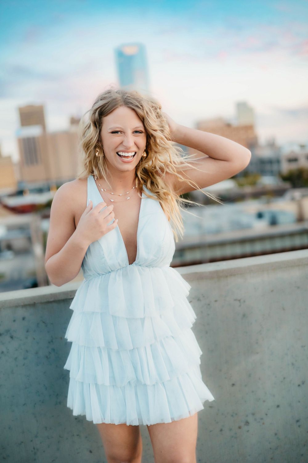 Smiling blonde woman in a white ruffled mini dress posing on a rooftop with a city skyline at sunset.