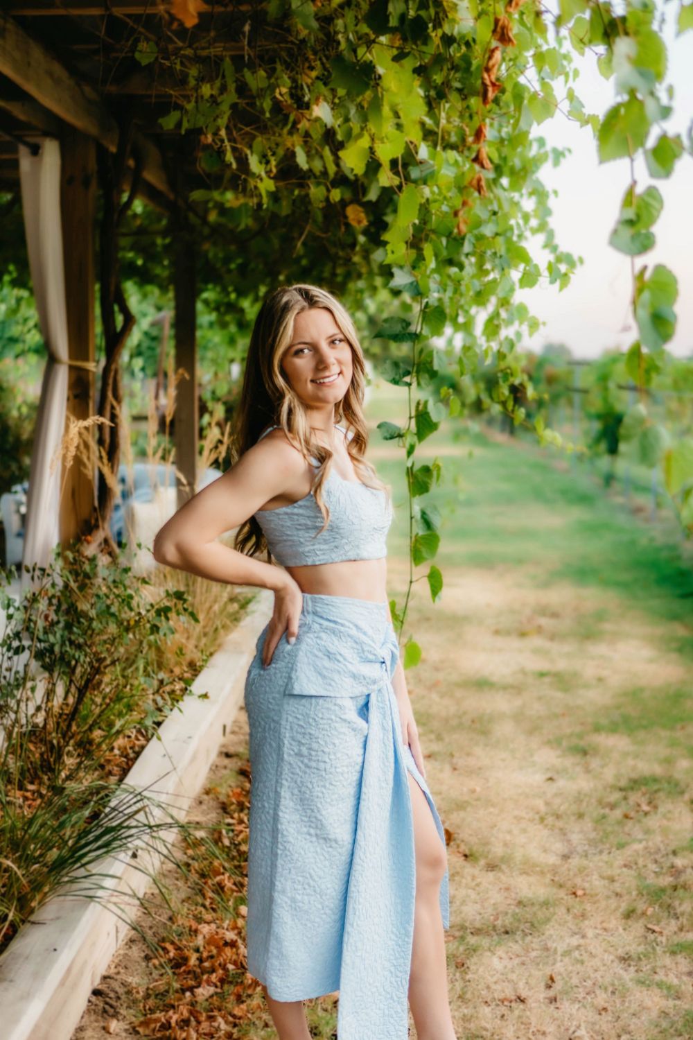 Smiling blonde woman in light blue crop top and midi skirt posing in a lush garden vineyard.