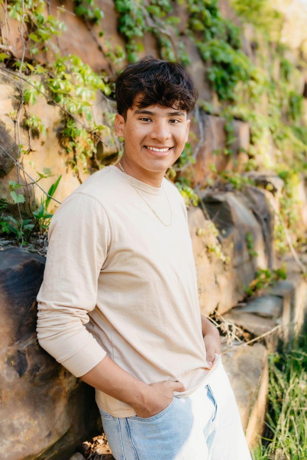 Smiling young man in beige long sleeve shirt and jeans leaning against a rocky wall with green ivy.