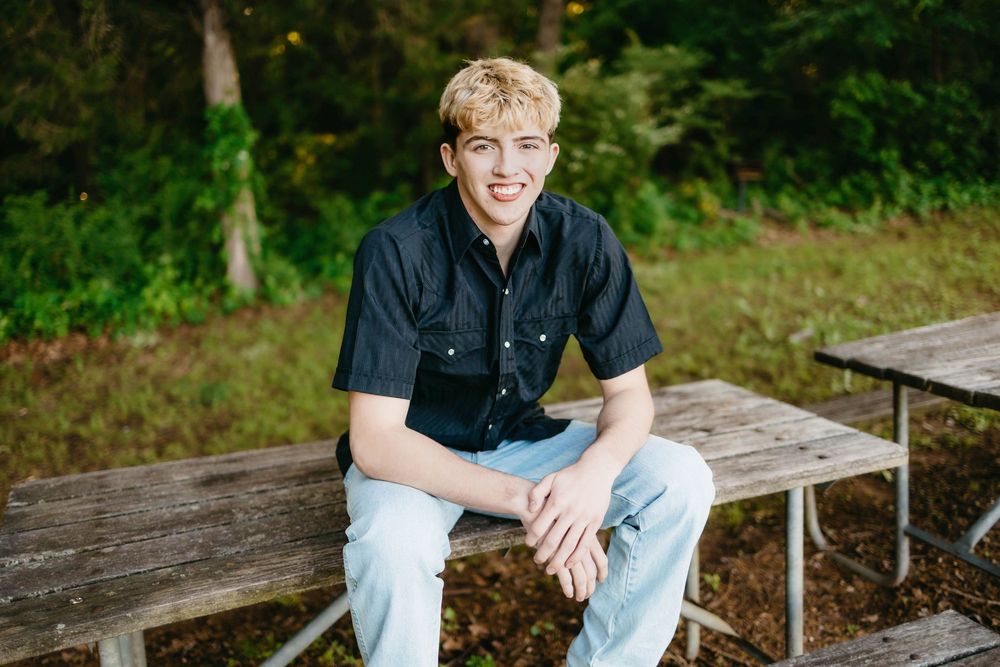 Smiling young man with blonde hair sitting on a picnic table outdoors wearing a black shirt and light jeans.