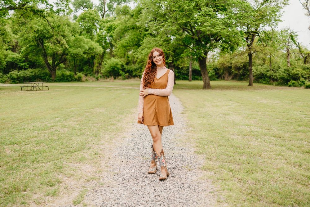 Young woman in tan dress and cowboy boots smiling on gravel path in lush green park.