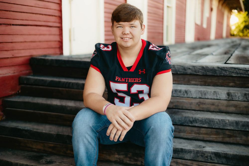 Teen boy in Panthers #53 football jersey sitting on wooden steps, smiling outdoors near red building.