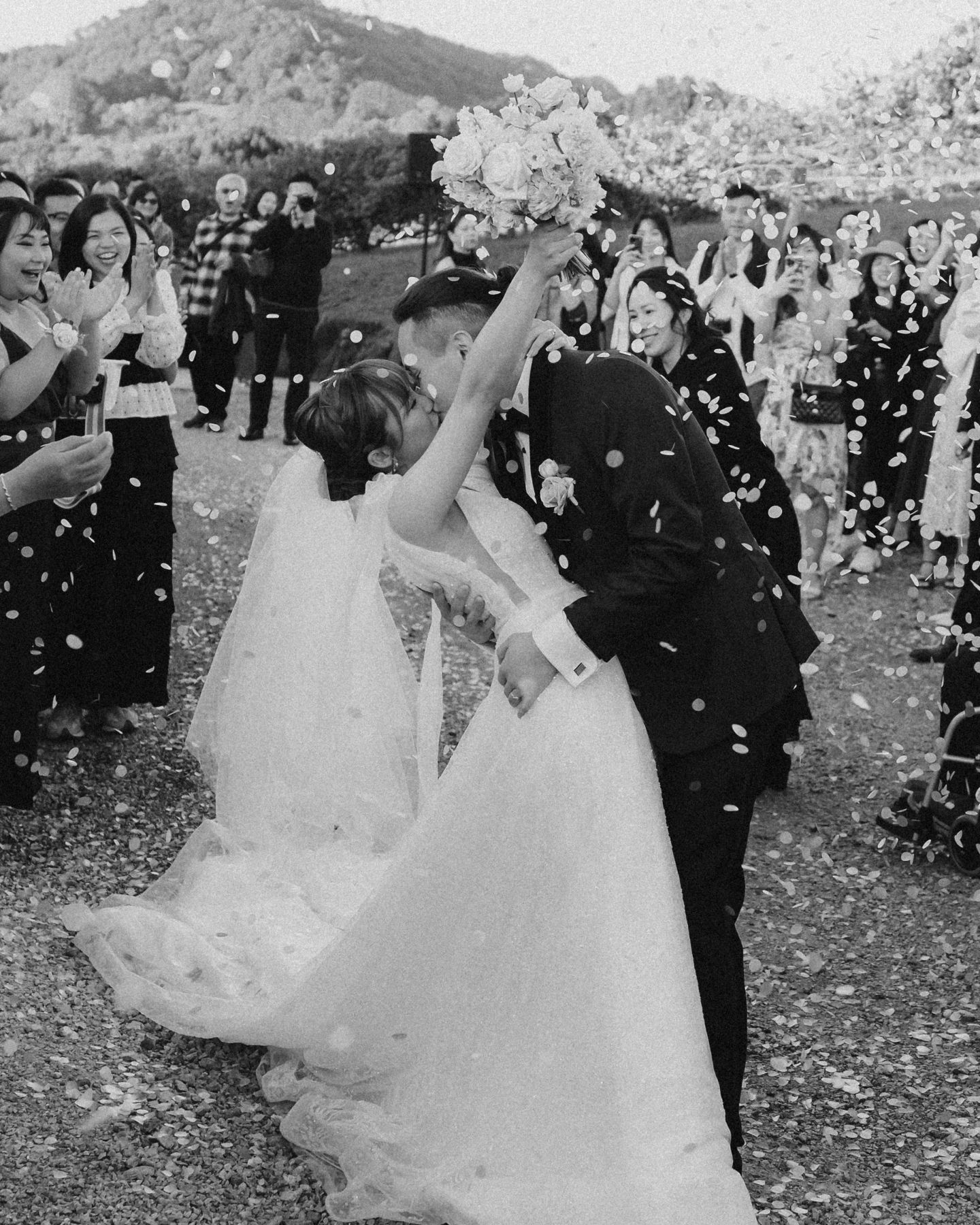 Black and white photo of newlyweds kissing amid confetti shower, surrounded by cheering guests at outdoor wedding ceremony.