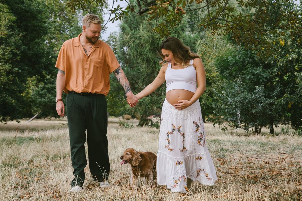Pregnant couple holding hands outdoors with a brown dog, surrounded by trees and golden grass.
