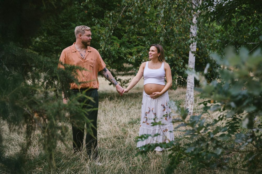 Pregnant couple holding hands in a lush green forest meadow, man in orange shirt, woman in floral skirt.