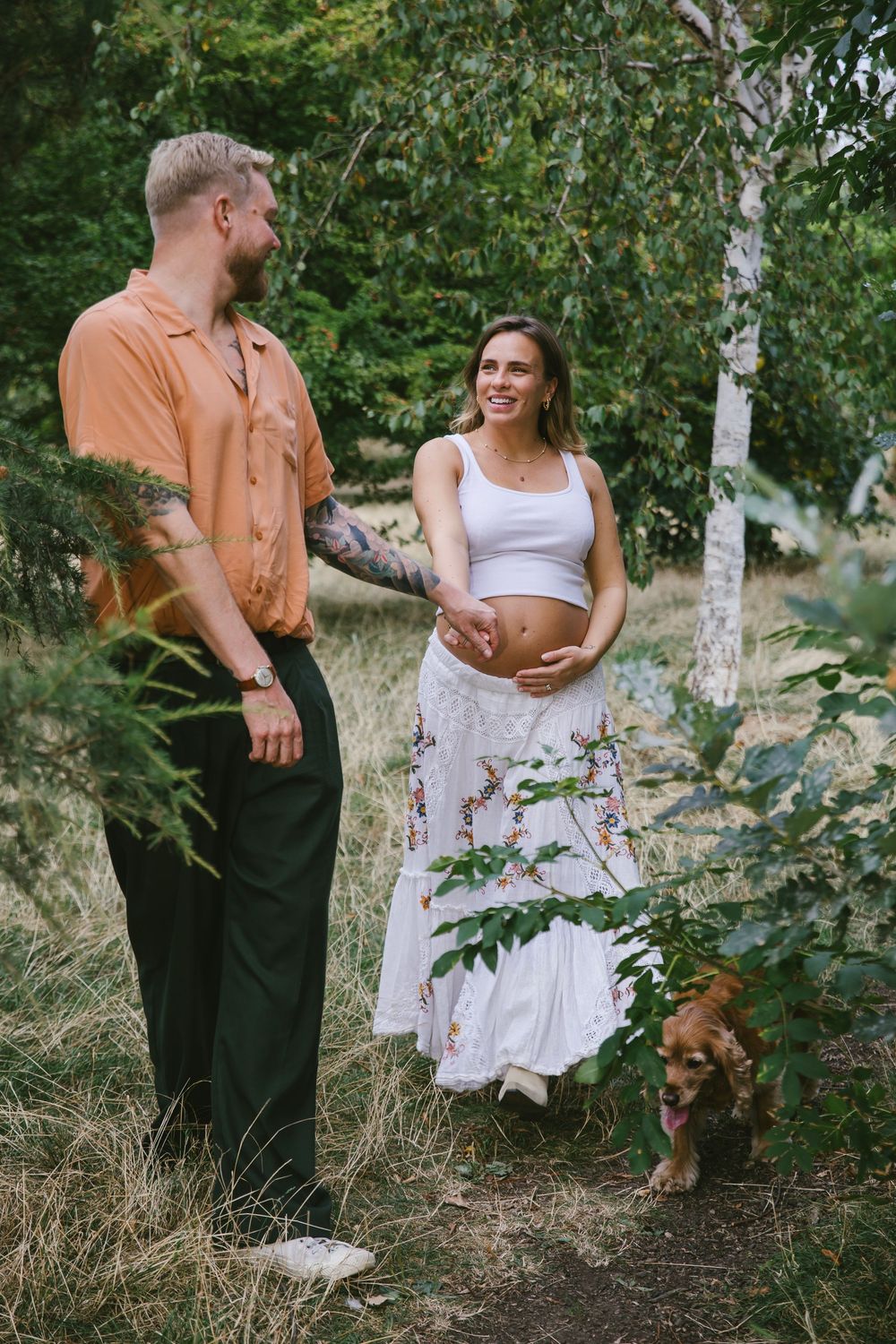 Smiling pregnant woman in white dress stands with tattooed man and dog in lush green forest setting.