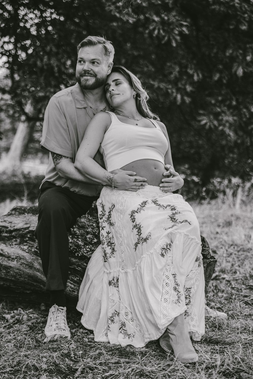 Black and white photo of a couple sitting on a log outdoors, woman in floral skirt cradling baby bump, maternity shoot.