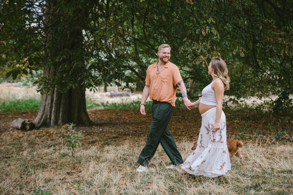 Couple walking hand-in-hand through a sunny park, man in orange shirt and green pants, pregnant woman in floral dress.
