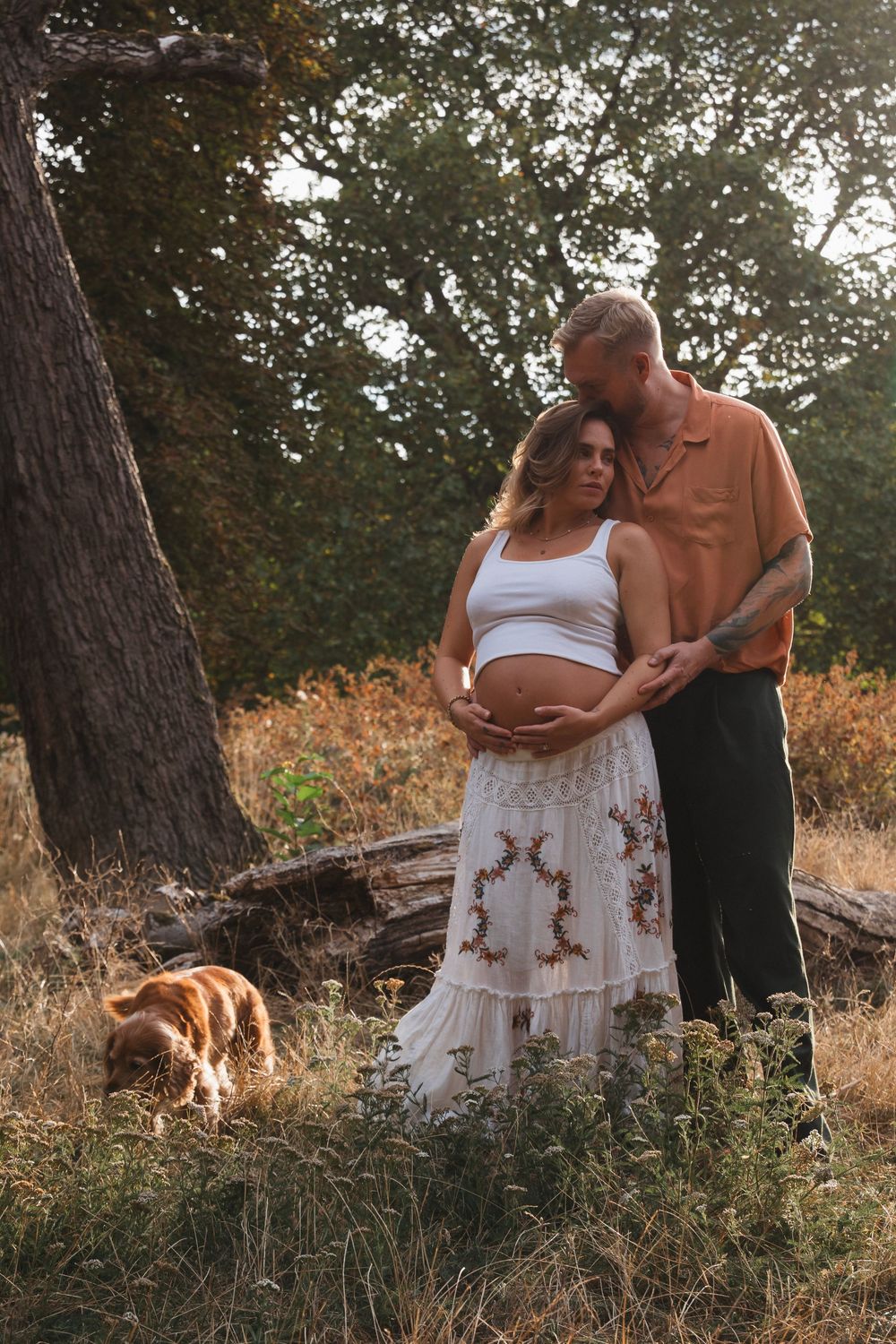 Couple tenderly embracing outdoors, pregnant woman in white crop top and floral skirt, golden retriever nearby in sunlit forest.