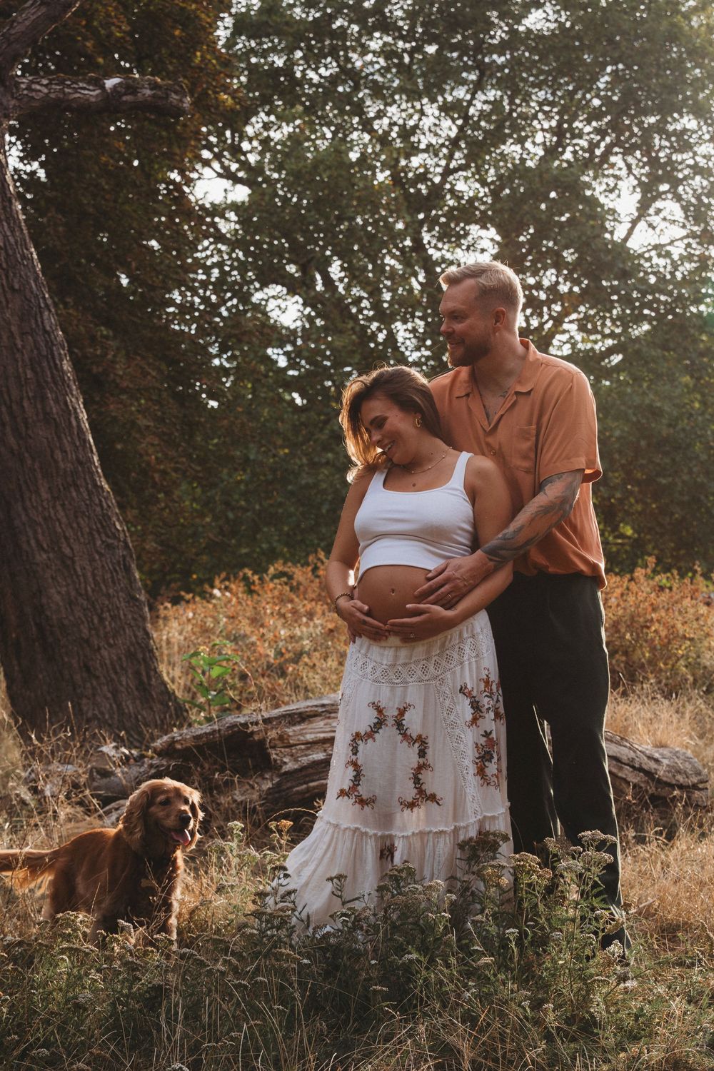 Couple embracing outdoors during golden hour, pregnant woman in white dress with partner and golden retriever dog.