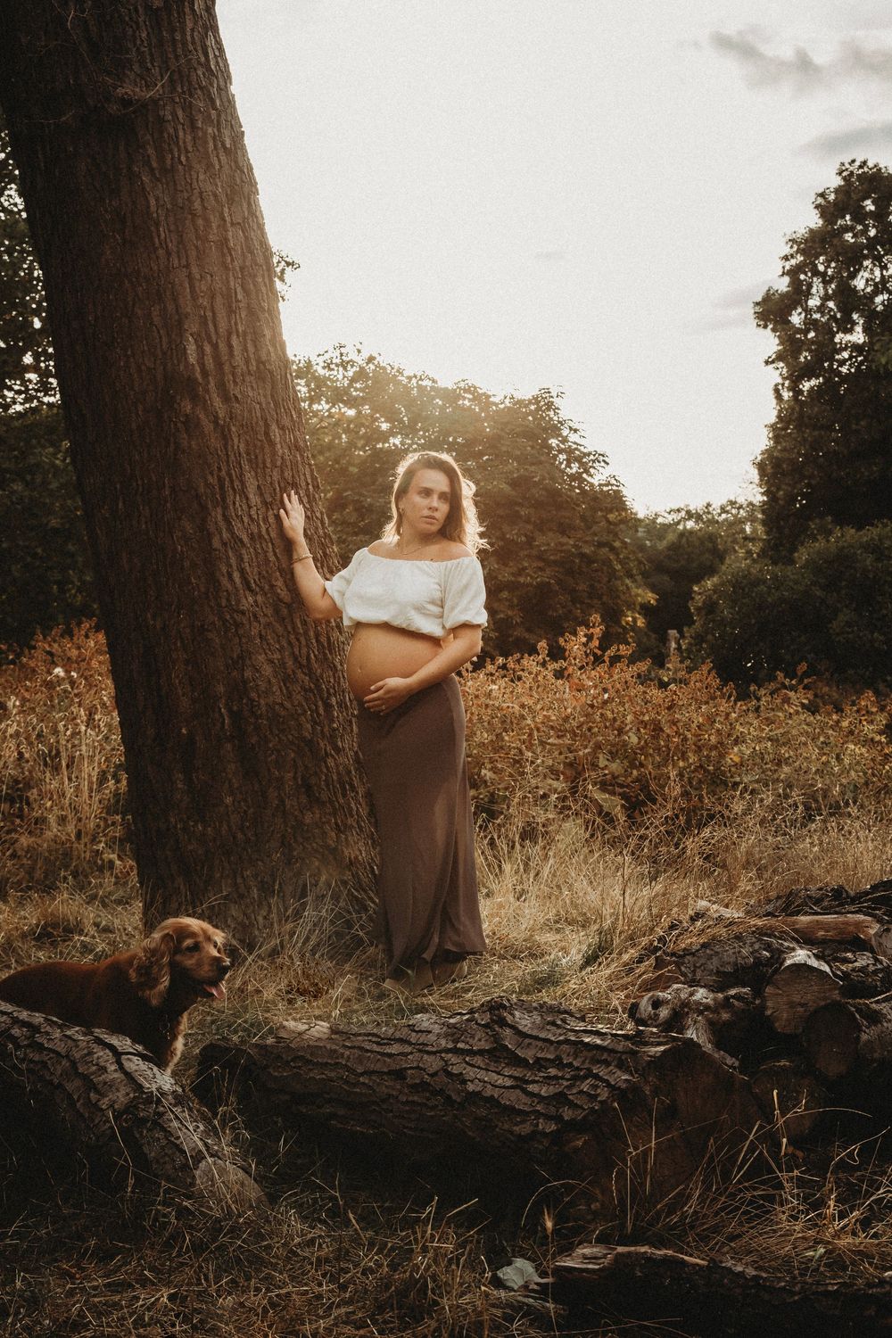 Pregnant woman in white top and brown skirt leaning against a tree outdoors with a dog nearby.