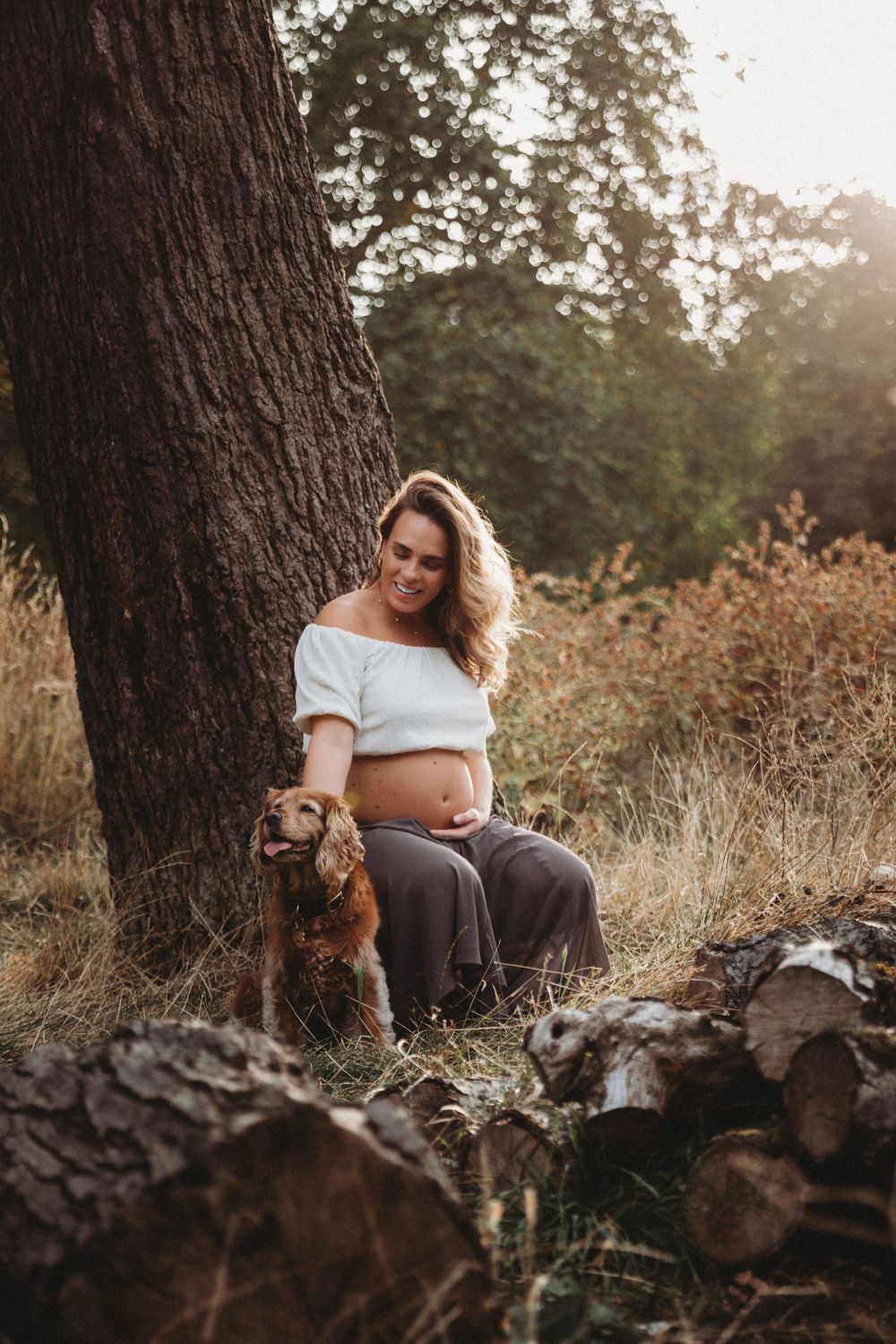 Smiling pregnant woman in white crop top sits against tree trunk with golden retriever dog in warm sunset forest setting.