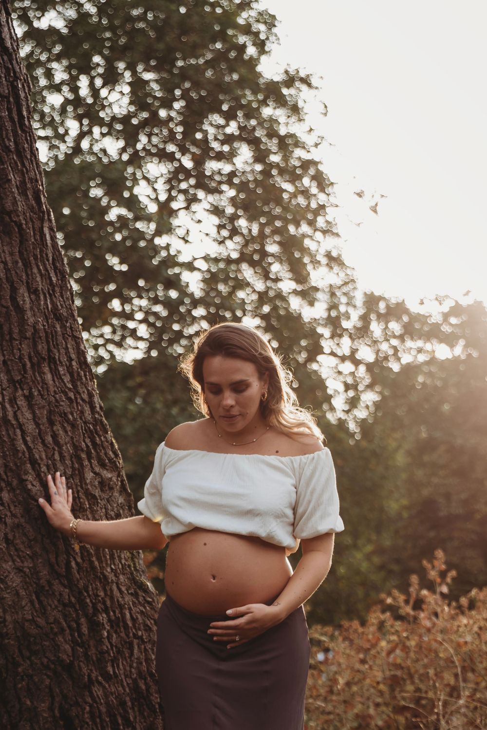 Pregnant woman in white off-shoulder top cradling her baby bump beside a tree in warm sunset light.
