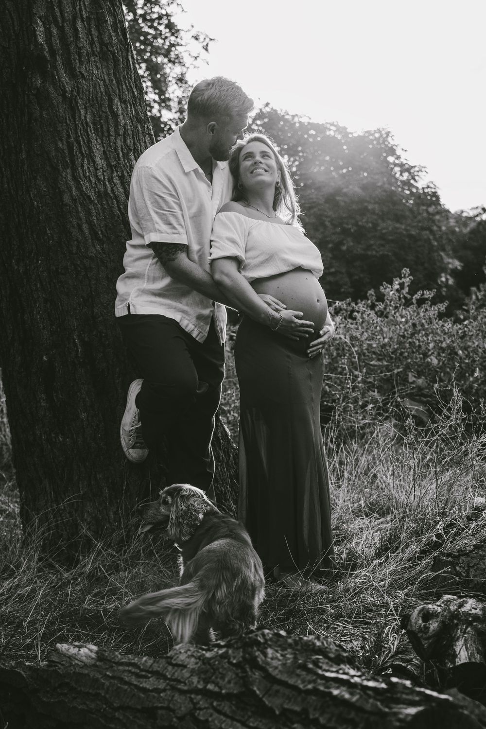 Black and white maternity photo of couple leaning against tree with dog, outdoors in natural setting.